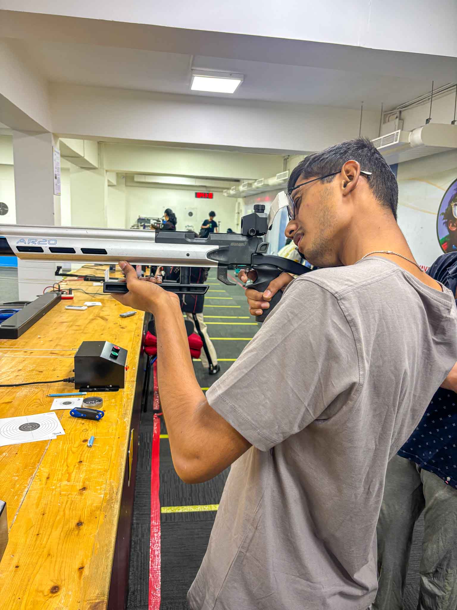 A shooter positioned in shooting stance aiming down the rifle barrel at an indoor shooting range.
